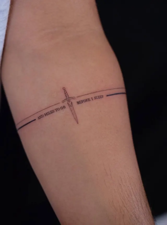 A forearm with a thin line armband and a tiny sword, with a line of fine lettering running around it
