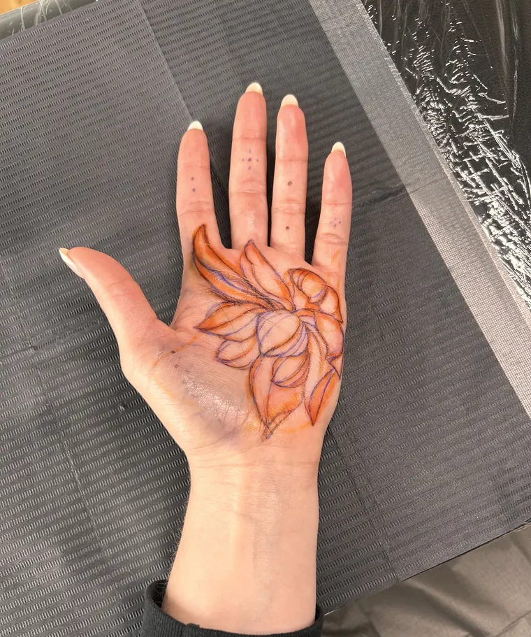 A palm resting on a grey paper drape with a lotus stencil drawn in orange and purple before tattooing