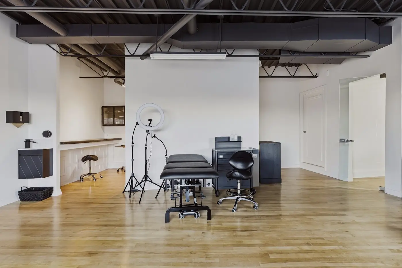 The studio: a bright white room with a black treatment table, ring light on a tripod, black stool and a maple hardwood floor